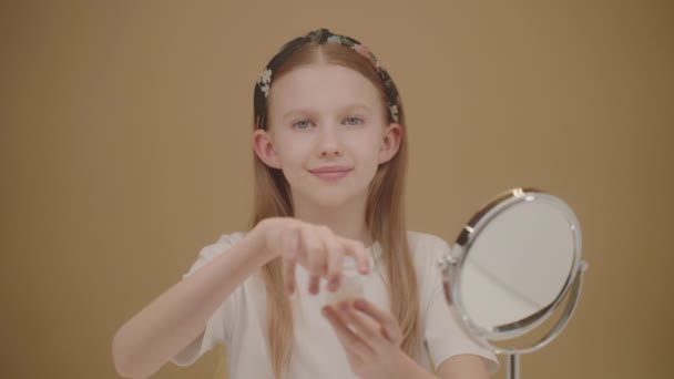 有趣的少女应用面霜 漂亮的金发姑娘看着相机 打开一瓶保湿霜 涂在她的脸颊上 青少年概念美容及护肤产品 — 图库视频影像