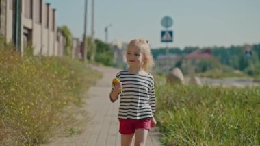 Küçük sarışın kız ağır çekimde güneşli bir günde yürüyor, sarı çiçekleri tutuyor, kameraya bakıyor ve gülümsüyor, mutlu anneler günü konsepti, eğlenceli yaz rüyası ve neşeli koşu.. 