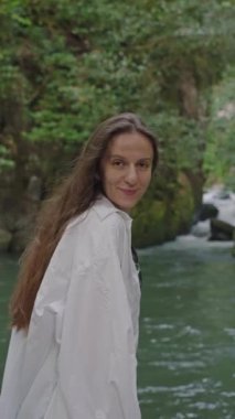 Dikey video. Mutlu stil sahibi, genç, sağlıklı esmer kadın gülüyor ve tropikal ormanda şelalenin yanında duran kameraya bakıyor. Dalgalı uzun saçlara dokunuyor. Ekoloji, yolculuk..