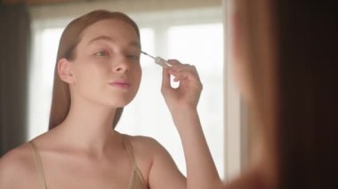 Güzel sarışın kadın aynaya bakarken makyaj fırçasıyla kirpiklerine jöle sürüyor. Maskara reklamı, kirpik uzatmaları, makyaj malzemeleri, cilt bakımı. Evde güzellik rutini