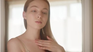 Ağır çekim. Güzel, ince, sarışın bir kız ve çıplak omuzları parmaklarıyla boynuna dokunuyor ve vücudunu bir aynanın önünde çeviriyor. Kadın vücut losyonu sürüyor ve boyun derisine masaj yapıyor. Vücut bakımı