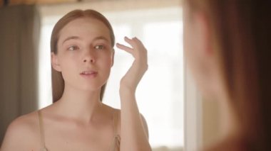 Temiz ciltli, alnına yakından dokunan, nemlendirici, serum süren ve aynaya bakan çekici sarışın bir kadın. Güzellik ve sağlık için cilt bakımı kavramı. Yavaş hareket. Ticari portre.