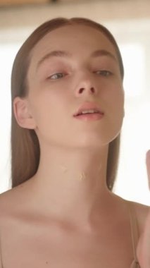 Dikey video. Güzel, ince, sarışın bir kız ve çıplak omuzları parmaklarıyla boynuna dokunuyor ve vücudunu bir aynanın önünde çeviriyor. Kadın vücut losyonu sürüyor, boyun derisine masaj yapıyor.. 