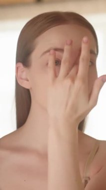 Dikey video. Mükemmel cilt tebessümü ve dokunaklı alnıyla genç, muhteşem sarışın kadın nemlendirici, serum ve aynaya bakıyor. Cilt bakım konsepti, sabah güzellik rutini..