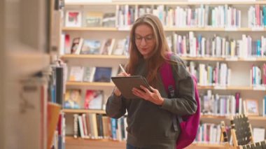 Tabletli ve sırt çantalı beyaz kadın modern kütüphanede kalemle işaretleme kitapları, fen projesinde çalışıyor, özel ders için ödev yapıyor. Kitap seçimi Çevrimiçi Eğitim Yüksek Öğrenim E-Öğrenim.