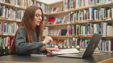 Ciddi derecede çekici beyaz kız öğrenci online öğrenim için dizüstü bilgisayar kullanıyor, kütüphanede sosyalleşiyor, öğretmenlik mesleki gelişimi, yüksek öğrenim, bir bilim projesi üzerinde çalışıyor.