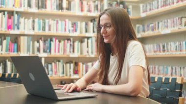Gözlüklü tatlı beyaz tenli esmer, kütüphanede öğrenci işi aramak için dizüstü bilgisayar kullanıyor, araştırma projesi için haber kaynaklarına kayıyor, sınav hazırlığı, yüksek öğrenim çevrimiçi, öğretmenler e-öğrenim