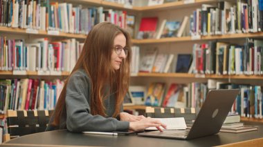 Beyaz tenli esmer kız gözlüklü öğrenci internetteki bir web sitesini izliyor, kalemle not alıyor, laptopta yabancı bir dil öğreniyor, uzaktan öğrenme kursuna ya da webcam öğretmenine göre