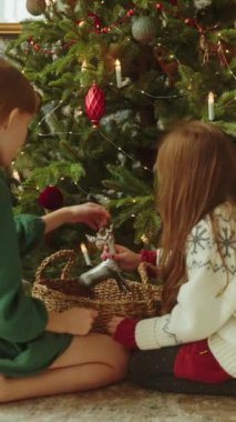 Dikey video. İki Kız Kardeş Noel ağacını birlikte süslüyor, Noel oyuncaklarını yavaş çekimde sepetten çıkarıyorlar, Mutlu Yıllar arifesi. Mutlu Noeller sinematik video koleksiyonu.