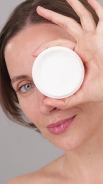 Dikey video: Çekici pozitif kadın kameraya özgüvenle bakıyor, elinde kozmetik krem kavanozuyla stüdyoda poz veriyor. Yüz bakımı, nemlendirici, parlayan cilt, spf güneş kremi reklamları..