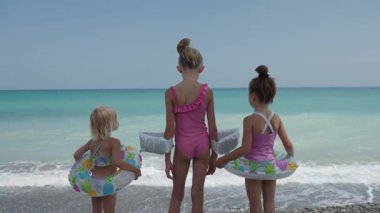 Mayolu üç kız çocuğu plajda yüzme yüzükleri tutuyor, denize bakıyor ve yüzmeye hazırlanıyorlar. Okyanus dalgaları, neşeli aile yaz tatilleri, tasasız çocukluk ve güvenli maceralar..