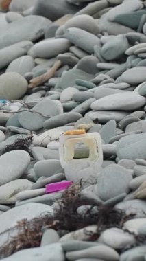 Dikey video: Şişeler ve ambalajlar da dahil olmak üzere plastik atık, çevresel kirliliği vurgulayarak kayalık bir plajı kirletiyor. Sahne, doğa ve insan etkisi arasındaki zıtlığı yakalar..