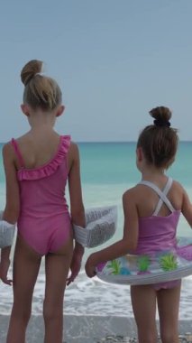 Dikey video: Mayolu iki kız çocuğu plajda yüzme yüzükleri tutuyor, denize bakıyor ve yüzmeye hazırlanıyorlar. Okyanus dalgaları, neşeli aile yaz tatilleri, tasasız çocukluk ve güvenli maceralar.