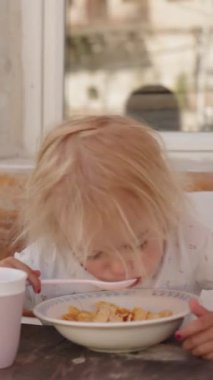 Dikey video: Bir yaz sabahı açık hava masasında buğday veya mısır gevreğiyle sağlıklı bir kahvaltı yapan komik küçük bir kızın yakın çekimi, gerçek hayattaki aile anları eğilimini yansıtıyor.