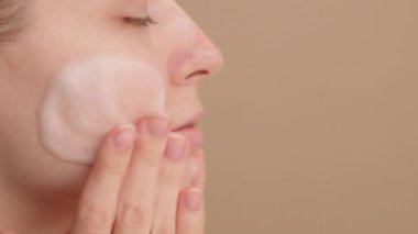 Yüzüne nazikçe temizleyici köpük süren bir kadının yakın çekimi, sağlıklı, ışıl ışıl bir cildi teşvik eden ve doğal güzelliğini artıran günlük cilt bakımını üstlenmesi. Hafif arkaplan. Boşluğu kopyala