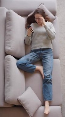 Dikey üst görünüm. Gözlüklü çekici beyaz kadın elinde akıllı bir telefonla kanepede yatıyor. İnternette sörf yapan neşeli genç bir kadın, web sörfü web sitesi, online alışveriş yapma kavramı.