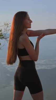 Dikey çekim. Uzun saçlı spor giyim mankeni güneş doğarken bir dağın tepesinde esneme egzersizi yapıyor, kadın vadiyi soluk kesici manzaranın keyfini çıkarıyor. Spor sutyen reklamları. 
