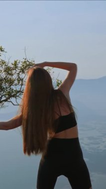 Dikey çekim tanınmaz halde. Uzun kahverengi saçlı, siyah spor sutyen ve şort giyen, dağ tepesinde gerinen genç bir kadın. Duruş düzeltmesi. Omurga bakımı. Spor elbiseli kadın.