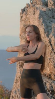 Dikey ekran. Siyah spor kıyafetleri giyen genç güzel bir kadın, canlı bir günbatımında, bir dağın tepesinde spor egzersizleri yapıyor, doğanın manzaralı güzelliğinin tadını çıkarıyor. Sırt esnekliği, iyi duruş