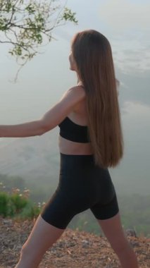 Dikey üst görünüm. Siyah spor sutyen ve şort giyen genç ve güzel bir kadın, arka planda sisli vadiyle birlikte dağ zirvesinde egzersiz yapıyor. Güzel duruş