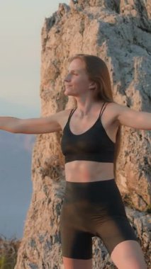 Dikey ekran. Siyah spor kıyafetleri giyen genç bir kadın, bir dağın tepesinde egzersiz yapıyor, spor salonunda temiz havanın tadını çıkarıyor. Omurga bakımı. Güzel duruş. Spor sutyen reklamları. 