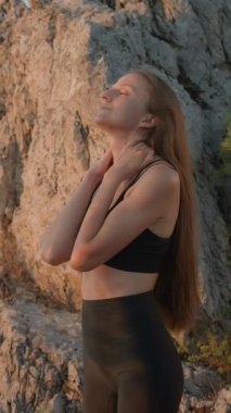 Dikey çekim. Uzun saçlı, siyah spor kıyafetleri giyen, kayaların yakınındaki bir spor seansından sonra boynuna dokunan genç bir sporcu. Kadın sporcu derin nefes almaya hazırlanıyor. Boyun masajı