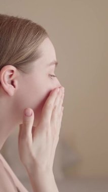 Dikey video: Sabah güzellik programı sırasında yüz serumu uygulayan genç bir kadının yakın çekimi. Minimalist, doğal cilt bakım konsepti gün ışığında filme alınır. Gen Z kendine özen, nazik yüz masajı..