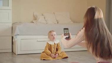 Anne, rahat bir evde, akıllı bir telefonla küçük kızının fotoğrafını çekiyor. Çocuk gülümsüyor ve poz veriyor. Çocukluk anılarını yakalamak, sevgi ve özen göstermek, duygusal aile bağları..