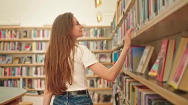 Kendine güvenen genç bir öğrenci, bir üniversite kütüphanesinde sakince kitaplara bakıyor, odaklanmış, neşeli bir ifadeyle, akademik keşif sürecinin tadını çıkarıyor. Modern bir kütüphanedeki kadın.