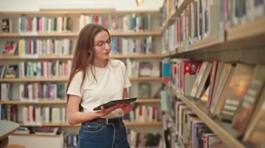 Üniversite öğrencisi kütüphanede sınavlara hazırlanıyor, elinde tabletle kitap arıyor. Akademik araştırma, kendi kendini yönetme, yüksek öğrenim. Modern bir kütüphanede genç bir kadın.