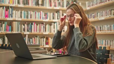 Sakin bir kız öğrenci bir üniversite kütüphanesinde dizüstü bilgisayarındaki bir derse katılır, kitaplıklar arasında özenle notlar alır, sınav hazırlığı ve mesafe öğrenmesine odaklanır. Harmanlanmış öğrenme.