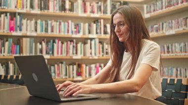 Üniversite kütüphanesinde dizüstü bilgisayarda yazan bir kız öğrenci, sınav sezonunda araştırmalarına odaklanmış, sessiz bir akademik ortamda kitap raflarıyla çevrili. Harmanlanmış öğrenme, eğitimdeki teknoloji.