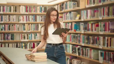 Üniversite öğrencisi kütüphanede kitap aramak için bir tablet kullanıyor. Modern akademik araştırma, proje araştırması, dijital araçlar ve sınav hazırlığı. Kadın akademik bilgi hedefini okuyor veya okuyor
