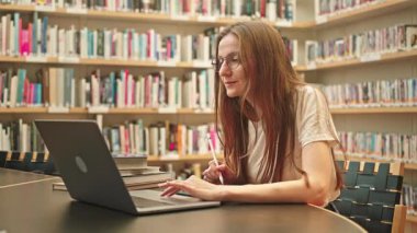 Üniversite veya halk kütüphanesinde dizüstü bilgisayar ve internet kullanan genç kız öğrenci, kitaplıklarla çevrili bir masada oturuyor, akademik araştırma yapıyor, notlar alıyor, çevrimiçi dersler dinliyor