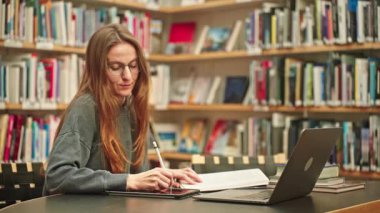 Gözlük okuyan mutlu ve odaklanmış bir kız öğrenci, online ders, araştırma projesi, webinar, sınav hazırlığı için bir üniversite kütüphanesinde notlar alıyor, bir dizüstü bilgisayar ve tablet kullanıyor..