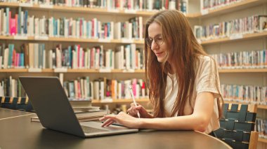 Üniversite kütüphanesinde dizüstü bilgisayar kullanan ve internette gezinen, etrafı kitaplıklarla çevrili bir masada oturan, öngörü araştırması yapan neşeli genç kız öğrenci notlar alıyor. Dijital öğrenme. 