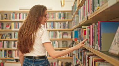 Üniversite kütüphanesinde duran kendine güvenen genç bir öğrenci sakince kitaplara bakıyor. Modern bir kütüphanedeki kadın. Kişisel gelişim. Öğrenci hayatı. Çalışma alışkanlıkları. Kişisel gelişim
