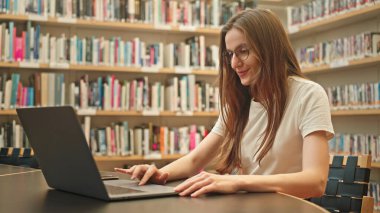 Üniversite kütüphanesinde dizüstü bilgisayarı olan bir kız öğrenci, sınav sezonunda araştırmalarına odaklanmış, etrafı sessiz bir akademik ortamda kitaplıklarla çevrili. Harmanlanmış öğrenme, eğitimdeki teknoloji.