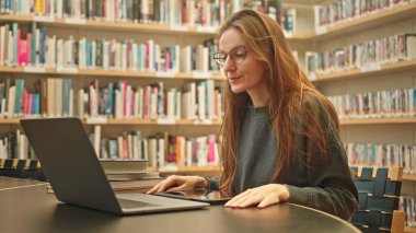 Sakin bir kız öğrenci bir üniversite kütüphanesinde dizüstü bilgisayarında bir derse katılıyor, kitaplıkların arasında, sınav hazırlıklarına, uzaktan öğrenmeye odaklanıyor. Harmanlanmış öğrenme.