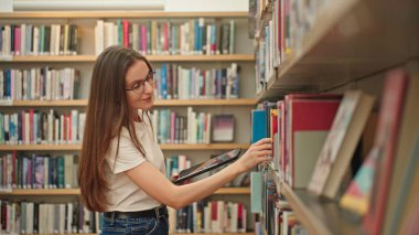 Üniversite öğrencisi kütüphanede sınavlara hazırlanıyor, elinde tabletle kitap arıyor. Akademik araştırma, kendi kendini yönetme, yüksek öğrenim. Modern bir kütüphanede genç bir kadın.