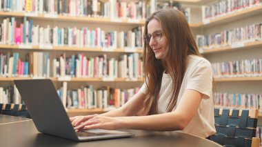 Üniversite kütüphanesinde dizüstü bilgisayarı olan bir kız öğrenci, sınav sezonunda araştırmalarına odaklanmış, etrafı sessiz bir akademik ortamda kitaplıklarla çevrili. Harmanlanmış öğrenme, eğitimdeki teknoloji.