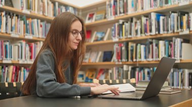 Sakin bir kız öğrenci bir üniversite kütüphanesinde dizüstü bilgisayarında bir derse katılıyor, kitaplıkların arasında, sınav hazırlıklarına, uzaktan öğrenmeye odaklanıyor. Harmanlanmış öğrenme. Akademik araştırma