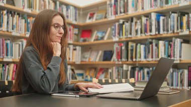 Sakin bir kız öğrenci bir üniversite kütüphanesinde dizüstü bilgisayarında bir derse katılıyor, kitaplıkların arasında, sınav hazırlıklarına, uzaktan öğrenmeye odaklanıyor. Harmanlanmış öğrenme. Akademik yazma