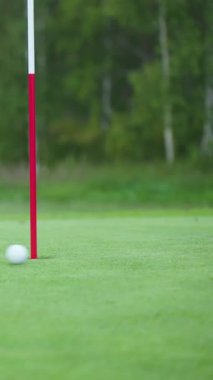 Beyaz golf topu yemyeşil yüzey boyunca pürüzsüzce yuvarlanıyor, parlak kırmızı ve beyaz bir iğneyle işaretlenmiş deliğe doğru ilerliyor, arka planda bulanık ağaçlar. Hedefi ıskaladın