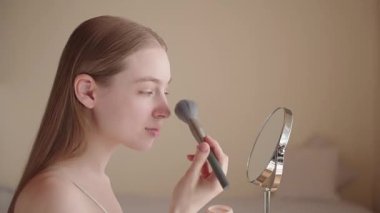 Neşeli genç bir kadın sabah güzellik rutininin bir parçası olarak fırçayla makyaj yapıyor. Mutlu bir Z Geni dişisi kendine bakmaktan ve güne hazırlanmaktan zevk alıyor. Doğal ışıklandırma, sıcak ve sakin atmosfer.