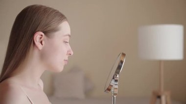 Genç bir kadın aynanın karşısında oturuyor, gülümsüyor ve cilt bakımının sonucuna hayran. Doğal görünüş, sağlıklı parlayan ten, gevşemiş ve tatmin olmuş ifade. Güzellik ve kişisel bakım konsepti.