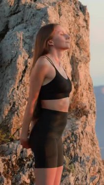 Spor kıyafetli genç bir kadın, gün batımında hafif bir gülümseme ile dışarıda esniyor ve nefes alıyor. Rahat duruş ve sakin enerji.