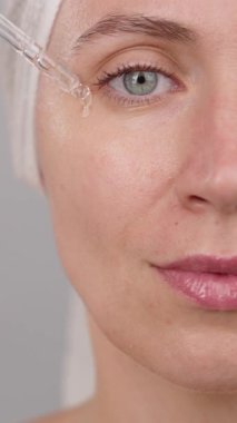 Yarım yüzlü, sakin bir kadının yakın plan portresi stüdyoda cildine serum süren bir kadın. Gri arka plan. Cilt bakımı ve güzellik kozmetiği için reklam konsepti, yüz serumu. Yavaş çekim.
