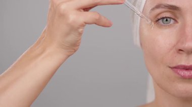 Yarı sakin kadın yüzünün yakın plan portresi. Stüdyoda cildine serum süren kadın. Gri arka plan. Cilt bakımı ve güzellik kozmetiği için reklam konsepti, yüz serumu. Uzayı kopyala Yavaş çekim.