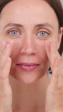 Dikey video. Güzel surat. Mavi gözlü, yüzüne dokunan esmer kadın kameraya bakıyor. Mükemmel cilde sahip güzel bir kadın model. Cilt reklamı konsepti, güzellik ürünleri